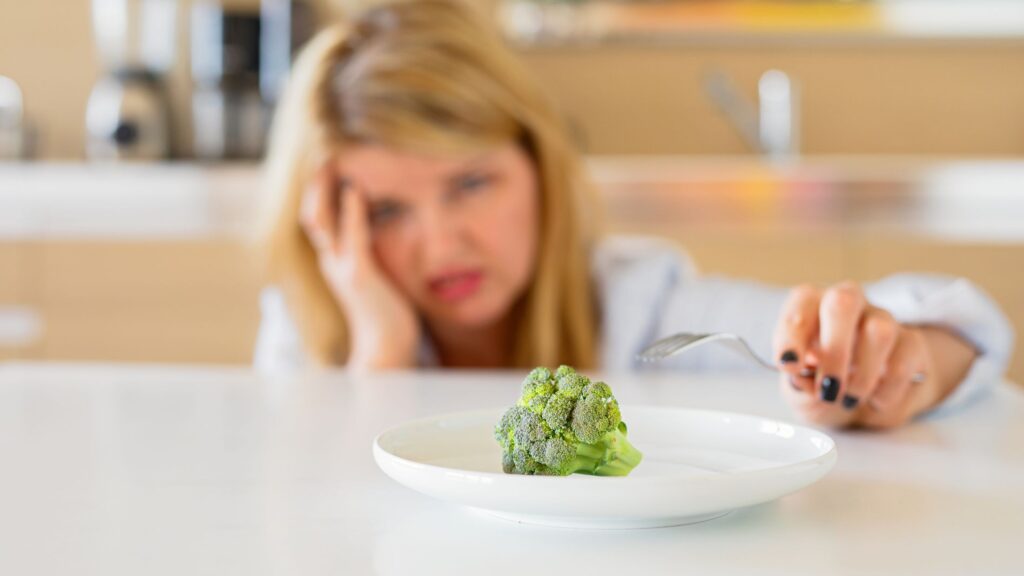 A woman looking at a piece of broccoli with hesitation, reflectant to adhere to an unhealthy restrictive diet.