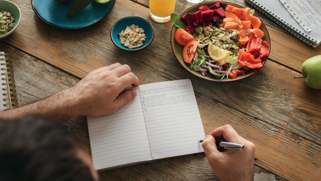 A person writing on an open notebook, beside a plate of wholesome food, a refreshing glass of orange juice, wholesome grains, and a crisp apple.