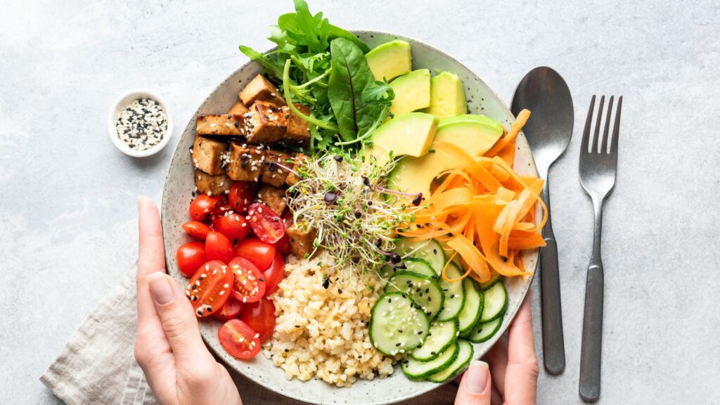 Two hands gently holding a plate filled with wholesome foods, including a fruits, vegetables, whole grains, and a protein source.