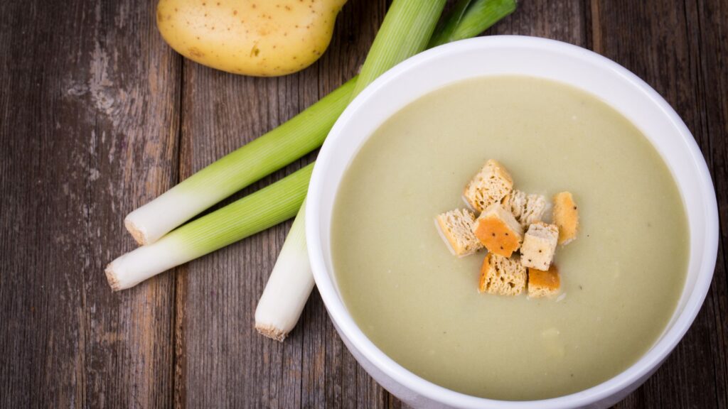 a bowl of leek soup with croutons along side with a potato and some leeks