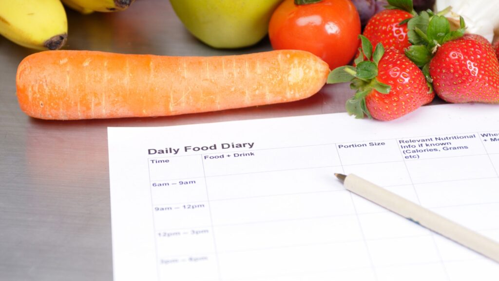 A sheet of paper displaying a daily food diary with detailed entries for each meal, including time, food and drink descriptions, portion sizes, and nutritional information, rests alongside a vibrant array of fresh produce, including carrots, strawberries, and a tomato.