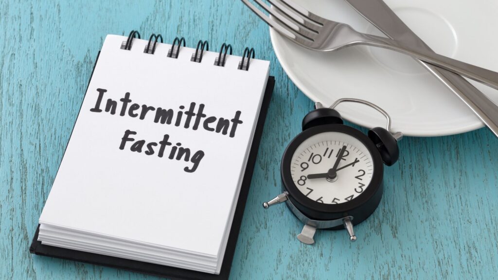 notebook with the words "intermittent fasting" written on the cover, lying on a wooden table next to a white alarm clock and an empty plate